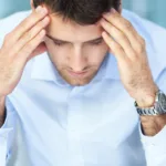 Man in light-blue button-down shirt with silver watch on left hand holds his head in his hands.