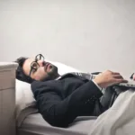 Man wearing a suit and glasses lies on his back in bed with a laptop open