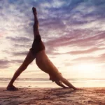 Man on beach has hands on sand and stretches his leg to the sky