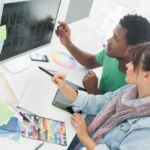 Woman with scarf and denim shirt pointing with a pen in hand sits next to man who points at monitor where they are both looking