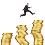 A man in a suit jumps between the middle stacks among five stacks of gold coins getting taller from left to right.