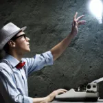 Man in blue button-down shirt and red bow tie wears a white fedora top hat and thick black glasses as he sits at a typewriter and reaches up toward a lightbulb floating in the air.