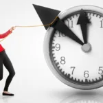 Blond woman in red blazer and black pants leaning back and pointing to large clock