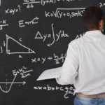 Man in white, long-sleeved shirt and jeans with a belt faces a blackboard where he writes formulas in chalk while holding a book