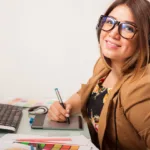 Girl in glasses wearing a brown blazer sits in front of computer with a sketch pad and color swatches in front of her