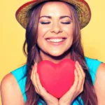 Smiling woman with long brown hair and straw hat wearing a blue sleeveless top holds a giant red heart