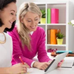 Two smiling women in business attire sit side-by-side while reviewing something on an iPad