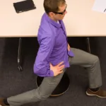 Man in grey slacks and purple top has hands on his hips and legs in a lunge position as he stretches in front of a desk.