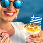 Woman in blue-tinted sunglasses smiles widely with red lips while holding a parfait dessert with a Greek flag in it.