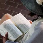 a girl wearing a black hat with a wide brim whose face we don't see is reading a book open on her lap