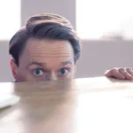 Preppy man with wide eyes peaking over top of table with hand on tap