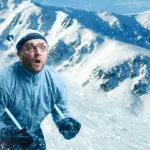 Man has ski poles tucked between arms with a terrified look on face while snow-covered mountains are in the background.