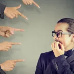 Man with glasses biting nails with multiple people pointing fingers at him.