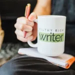 Close-up of hand holding a pen and also gripping white mug sitting on top of a spiral-bound notebook with the words "Filthy Rich Writer" on the mug.