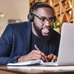 Black man in glasses, headphones, and blue blazer is in a cafe and eagerly writing down information while looking at his laptop.