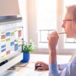 A woman wearing glasses and a blue button-down top with hair tied back in a clip looks at a desktop computer screen showing a calendar while one hand hovers on her mouse and another holds a pen to her mouth.