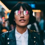 Woman with cropped haircut and leather jacket with white button-down top walks through a city, whose lights are reflected in her aviator glasses.