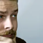 Close-up of man's face with hand curved around chin in thought and eyebrow raised.