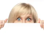 Blond woman with blue eyes peers over the top of a blank white page she holds in front of her nose