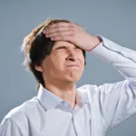 Brown-haired white man in a light blue button-down shirt has his eyes closed and his hand slapped against his forehead.