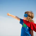 Young boy wearing a mask and a red cape pumps his fist and straight arm into the arm