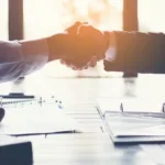 Two arms reach out shaking hands across a table with papers scattered on it and a burst of light in the background.