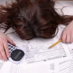 Woman is face down, head in a pile of tax documents with a calculator in one hand and a pencil in the other.