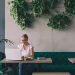 Woman with blonde hair and white shirt sits at a cafe desk typing on her white laptop with greenery sprawling behind her.