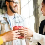 Young woman receives a red box with red bow from a colleague in yellow sweater as a bearded man in glasses looks on smiling.