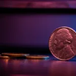 A nickel stands up on its edge with several nickels sitting on the table beside it while a deep violet color takes up the background.