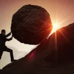 Man in suit is silhouetted against a setting sun as he scales a mountainside, pushing a large boulder up the mountain.