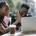 Young Black couple sitting at picnic table with laptop and coffee cup high fives.