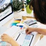 Woman sits at a desk creating her online portfolio with papers strewn on either side of the keyboard.